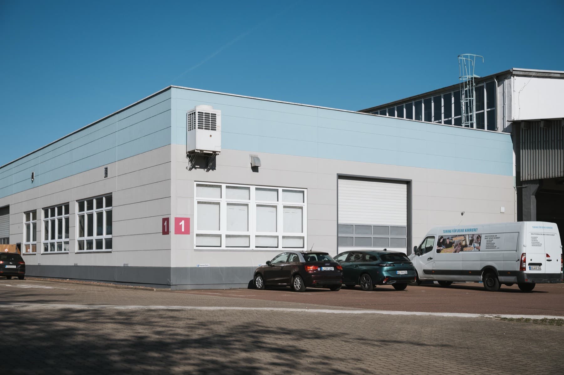Halle 1 — Außenansicht der Gebäudeecke mit Hallennummer, Fensterband, Rolltor und Parkplätzen davor
