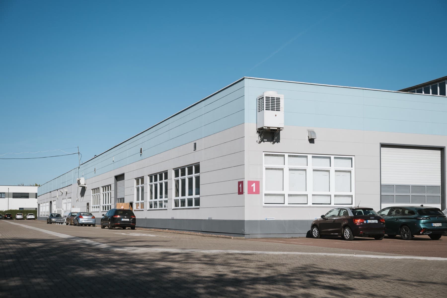 Halle 1 — Außenansicht entlang der hellgrauen Fassade mit Fensterreihen und Parkplätzen an der Zufahrtsstraße