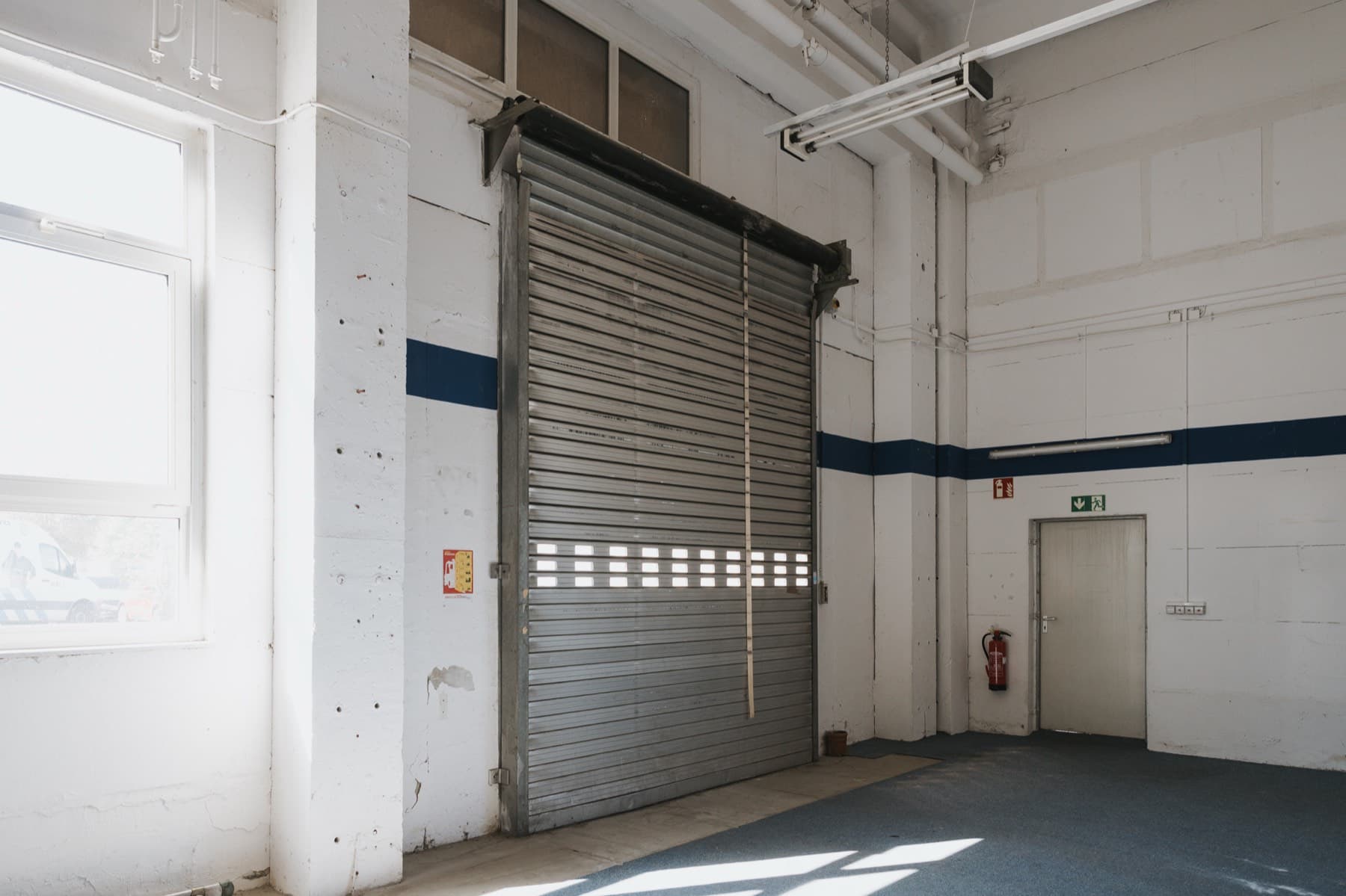 Halle 1 — Innenansicht mit Rolltor, Nebentür und Feuerlöscher an weißer Wand mit blauem Streifen