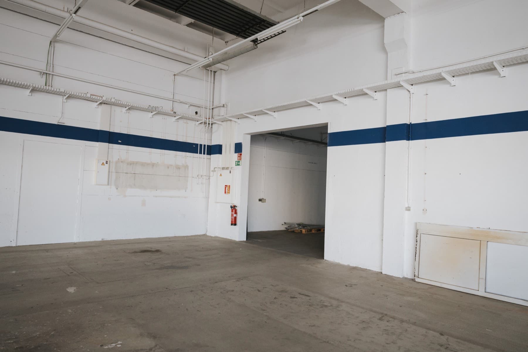 Halle 1 — leere Hallenfläche mit Betonboden, blauem Wandstreifen und breitem Durchgang zum Nebenraum