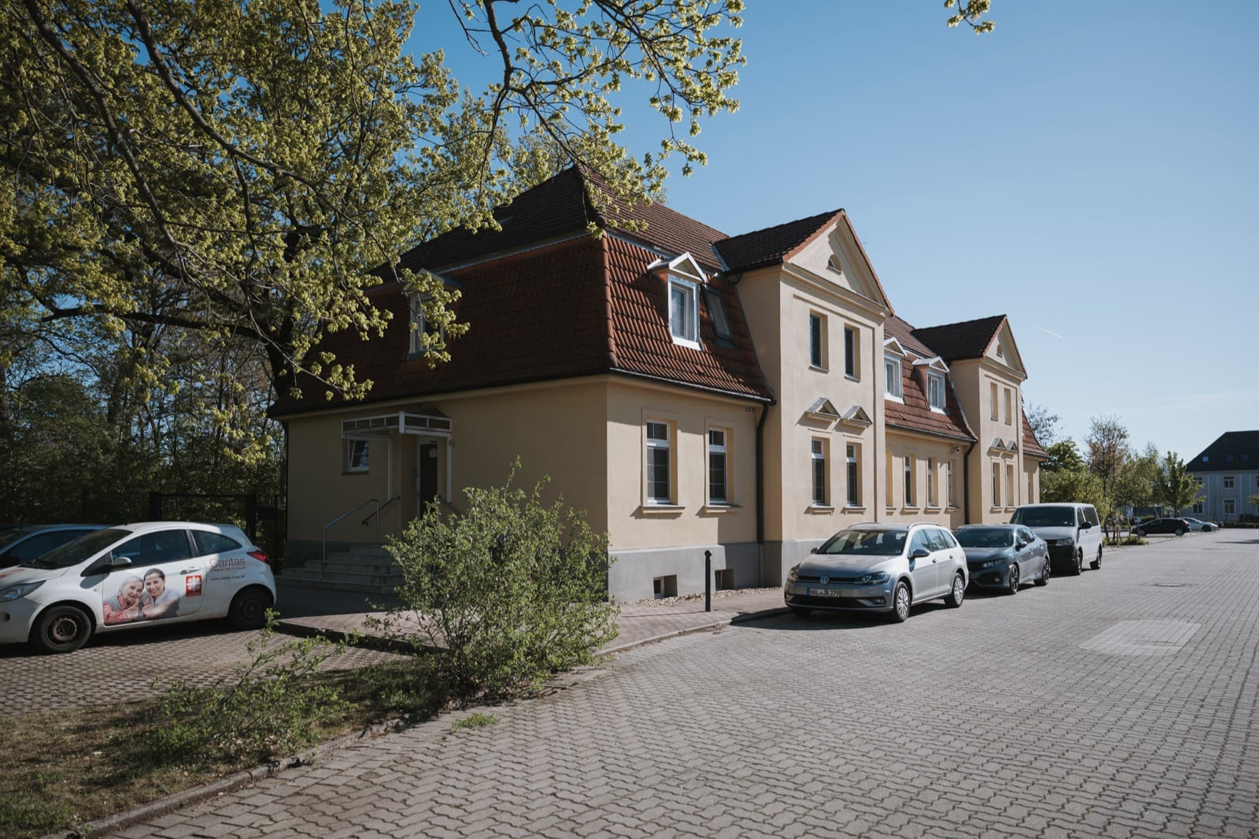Halle 11 — Straßenansicht des gelben Altbaus mit Mansarddach und Eingangstreppe unter Bäumen