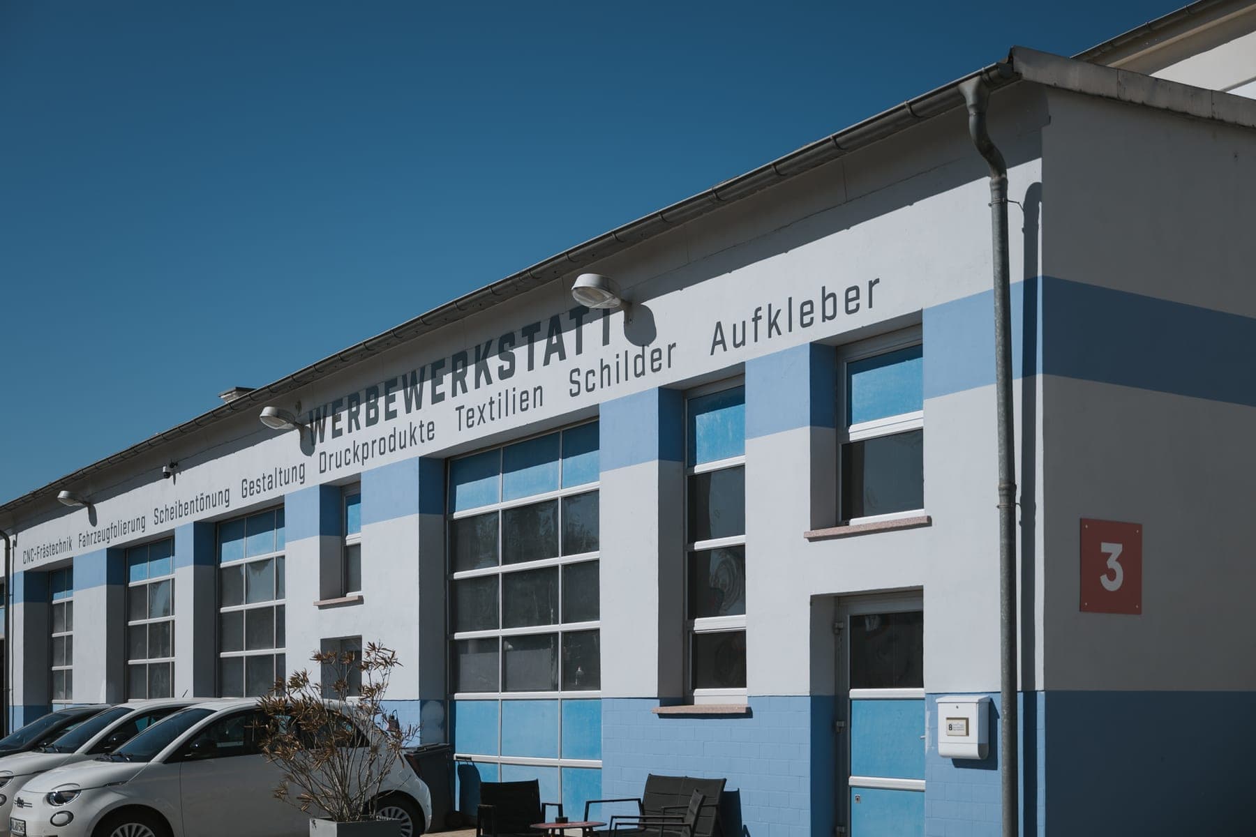 Blau-weiß gestreifte Fassade der Halle 3 mit Werbewerkstatt-Schriftzug über großen Sprossenfenstern bei Sonnenschein