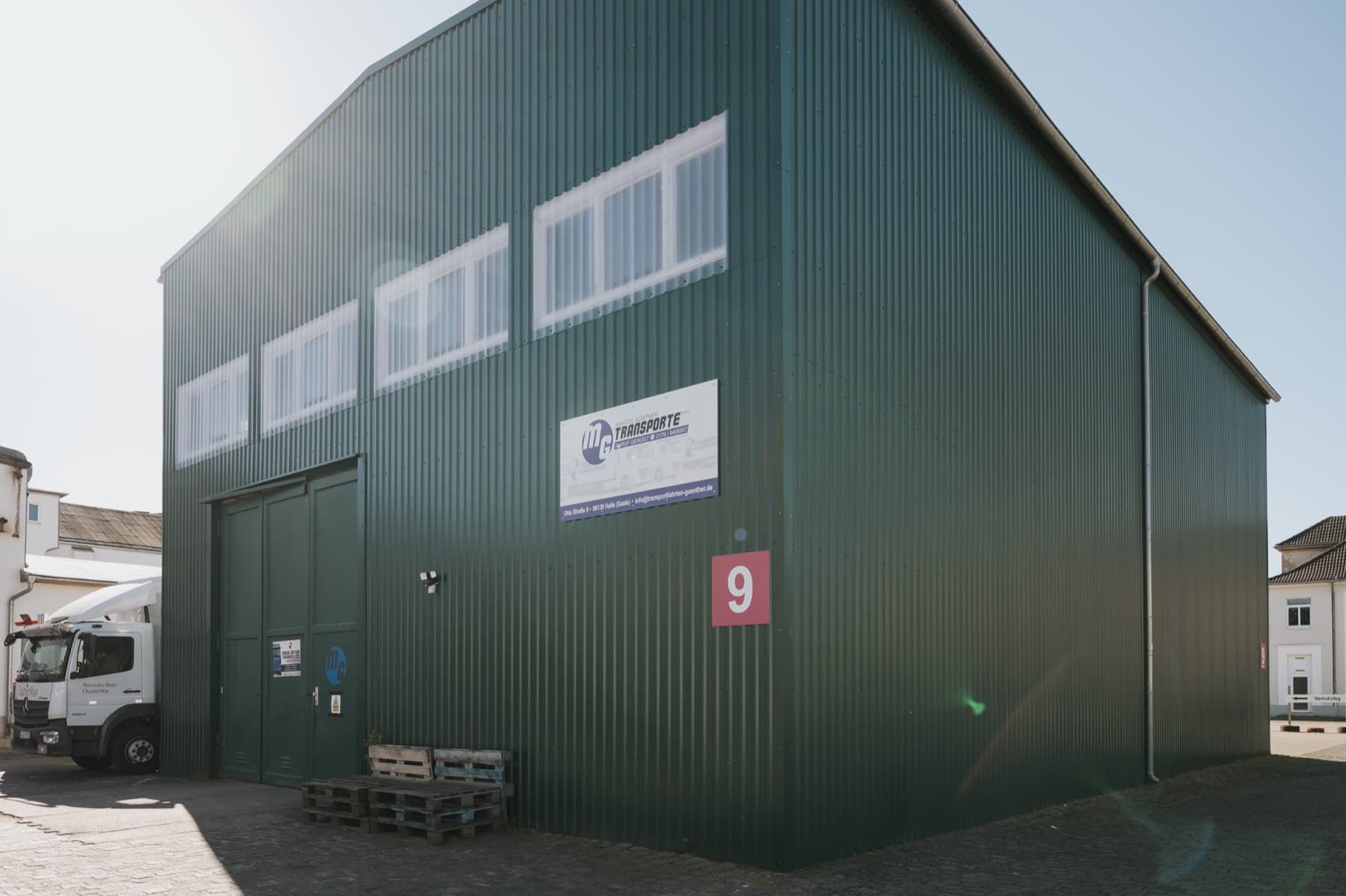 Halle 9 — grüne Wellblechhalle mit Einfahrtstor, Firmenschild und Lkw vor dem Eingang